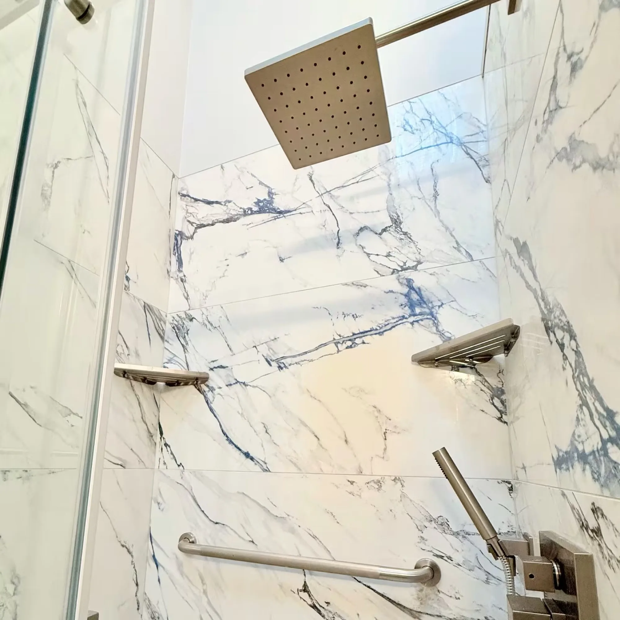 Shower interior in book-matched white marble-look tile with a square rain head and chrome grab bar